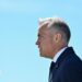 Mark Carney stands in profile, facing to the left, against a clear light blue sky. He wears a dark blue suit and tie.