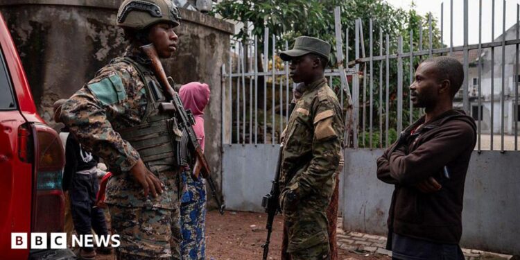 Two men dressed in military uniform and holding rifles stand alongside a member of the public in a brown hoody and trousers. A woman in a pink hoody has her back to the camera in the background