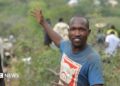 Manasse Lomachar in a blue T-shirt with a cartoon pig printed on it looks at the camera but gestures behind him with his right hand. In the background, out of focus, people can be seen working among the shrubs.