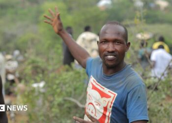 Manasse Lomachar in a blue T-shirt with a cartoon pig printed on it looks at the camera but gestures behind him with his right hand. In the background, out of focus, people can be seen working among the shrubs.