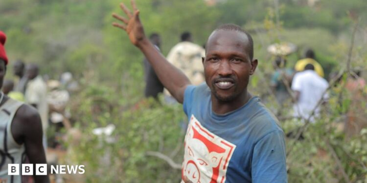 Manasse Lomachar in a blue T-shirt with a cartoon pig printed on it looks at the camera but gestures behind him with his right hand. In the background, out of focus, people can be seen working among the shrubs.