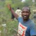 Manasse Lomachar in a blue T-shirt with a cartoon pig printed on it looks at the camera but gestures behind him with his right hand. In the background, out of focus, people can be seen working among the shrubs.