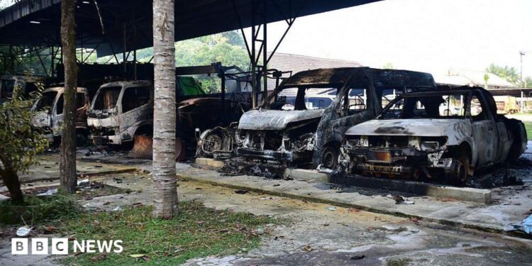 Five burned out vehicles are lined up under a tin roof structure