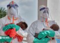 Two health workers in PPE each hold a young baby.