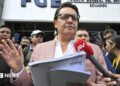 Fernando Villavicencio speaks into a microphone while holding papers and gesturing with one hand on the steps of a building surrounded by people. He wears a pink jacket, white shirt and lack-rimmed glasses.