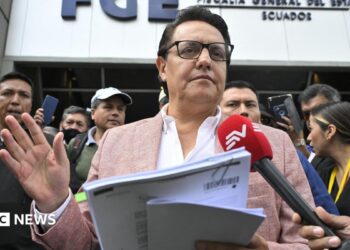 Fernando Villavicencio speaks into a microphone while holding papers and gesturing with one hand on the steps of a building surrounded by people. He wears a pink jacket, white shirt and lack-rimmed glasses.