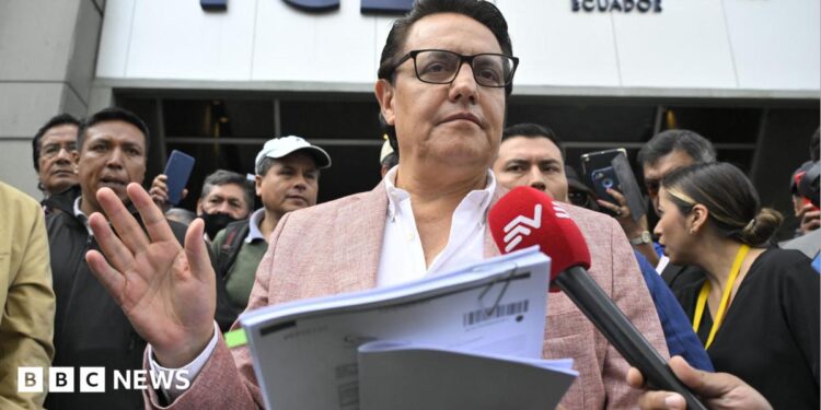 Fernando Villavicencio speaks into a microphone while holding papers and gesturing with one hand on the steps of a building surrounded by people. He wears a pink jacket, white shirt and lack-rimmed glasses.