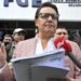 Fernando Villavicencio speaks into a microphone while holding papers and gesturing with one hand on the steps of a building surrounded by people. He wears a pink jacket, white shirt and lack-rimmed glasses.