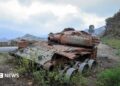 A rusted, wrecked tank lies on the side of a rural road. Mountains are visible in the background.