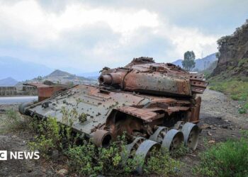 A rusted, wrecked tank lies on the side of a rural road. Mountains are visible in the background.