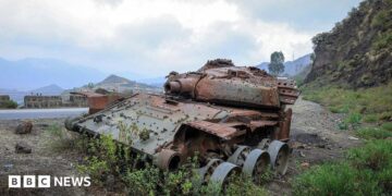 A rusted, wrecked tank lies on the side of a rural road. Mountains are visible in the background.