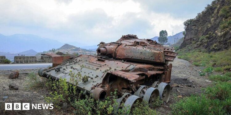 A rusted, wrecked tank lies on the side of a rural road. Mountains are visible in the background.