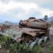 A rusted, wrecked tank lies on the side of a rural road. Mountains are visible in the background.