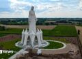 A statue of the Virgin Mary rises out of green and yellow fields in Poland on a cloudy sky.