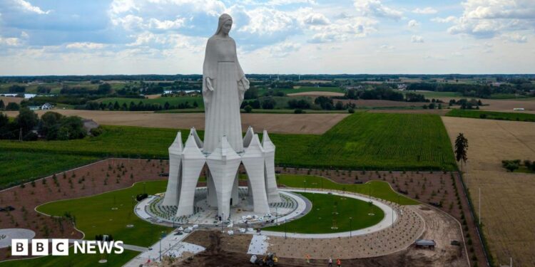 A statue of the Virgin Mary rises out of green and yellow fields in Poland on a cloudy sky.
