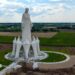 A statue of the Virgin Mary rises out of green and yellow fields in Poland on a cloudy sky.