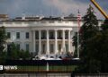 Exterior shot of the White House's South lawn, the Truman balcony (portico area enclosed by six white columns in the Ionic style, with a distinctive scroll-shaped ornaments called volutes on the capital. A yellow crane can be seen emerging from trees to the left of the frame