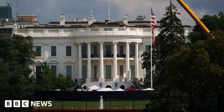 Exterior shot of the White House's South lawn, the Truman balcony (portico area enclosed by six white columns in the Ionic style, with a distinctive scroll-shaped ornaments called volutes on the capital. A yellow crane can be seen emerging from trees to the left of the frame
