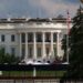 Exterior shot of the White House's South lawn, the Truman balcony (portico area enclosed by six white columns in the Ionic style, with a distinctive scroll-shaped ornaments called volutes on the capital. A yellow crane can be seen emerging from trees to the left of the frame