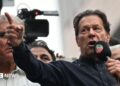 Imran Khan wearing a black button-up shirt and speaking into a microphone, points dramatically. Behind him are other men, and a television camera