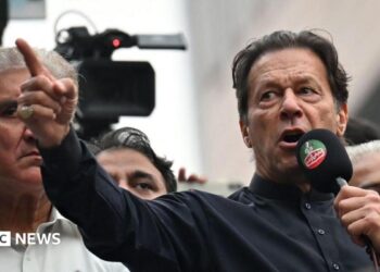 Imran Khan wearing a black button-up shirt and speaking into a microphone, points dramatically. Behind him are other men, and a television camera