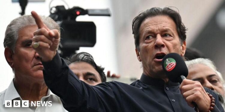 Imran Khan wearing a black button-up shirt and speaking into a microphone, points dramatically. Behind him are other men, and a television camera