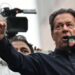 Imran Khan wearing a black button-up shirt and speaking into a microphone, points dramatically. Behind him are other men, and a television camera