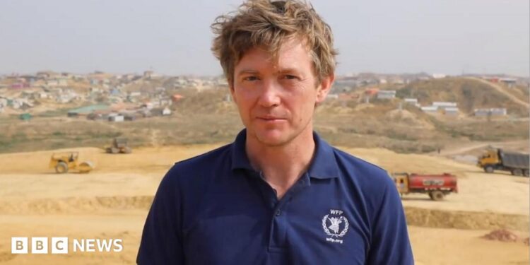 A man with brown hair looking at the camera, he is wearing a navy polo top