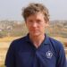 A man with brown hair looking at the camera, he is wearing a navy polo top