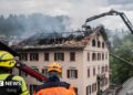 Large flames come out of the windows in the top floor of a building at night.