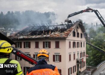 Large flames come out of the windows in the top floor of a building at night.