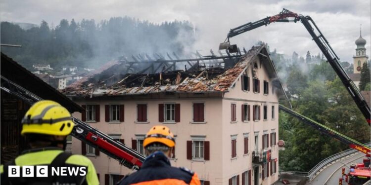 Large flames come out of the windows in the top floor of a building at night.