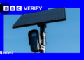 A black Flock surveillance camera attached to a pole with a solar panel attached above it