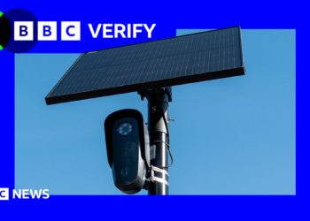 A black Flock surveillance camera attached to a pole with a solar panel attached above it