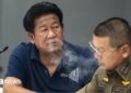 Former lawmaker Chalong Riewrang in a blue t-shirt exhales out cigarette smoke at a police station where he was allowed to hold a press conference after allegedly shooting and killing former police officer Thongchai Yenprasert. A police officer in uniform and glasses is seated to his right.