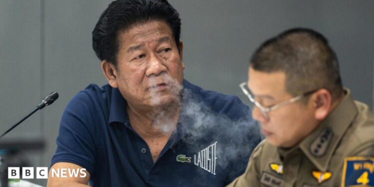 Former lawmaker Chalong Riewrang in a blue t-shirt exhales out cigarette smoke at a police station where he was allowed to hold a press conference after allegedly shooting and killing former police officer Thongchai Yenprasert. A police officer in uniform and glasses is seated to his right.