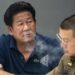 Former lawmaker Chalong Riewrang in a blue t-shirt exhales out cigarette smoke at a police station where he was allowed to hold a press conference after allegedly shooting and killing former police officer Thongchai Yenprasert. A police officer in uniform and glasses is seated to his right.