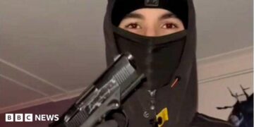 A teenager wearing a black balaclava and hoodie holding up a handgun, with his gloved hand on the trigger