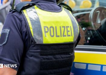 A German police officer standing next to a police vehicle. The officer wears an operational vest that reads "polizei".