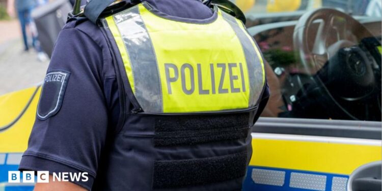 A German police officer standing next to a police vehicle. The officer wears an operational vest that reads "polizei".