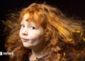 Close-up portrait of a young child with long, curly red hair flowing outward as if caught by a breeze. The child is turned slightly to one side and looking toward the camera, wearing a dark top against a dark, softly blurred background. Warm lighting highlights the texture and movement of the hair.