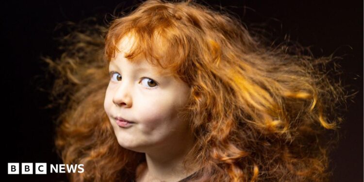 Close-up portrait of a young child with long, curly red hair flowing outward as if caught by a breeze. The child is turned slightly to one side and looking toward the camera, wearing a dark top against a dark, softly blurred background. Warm lighting highlights the texture and movement of the hair.