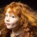 Close-up portrait of a young child with long, curly red hair flowing outward as if caught by a breeze. The child is turned slightly to one side and looking toward the camera, wearing a dark top against a dark, softly blurred background. Warm lighting highlights the texture and movement of the hair.