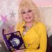 Dolly Parton sits holding a purple children's book with her pink fingernails, wearing a bright yellow suit and matching chunky jewelry against a childlike backdrop with stars and a googly-eyed pillow
