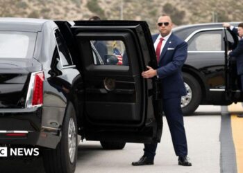 Two US Secret Service agents, both in suits, one in sunglasses, hold open the back passenger doors on two versions of the "Beast" armoured limousine.