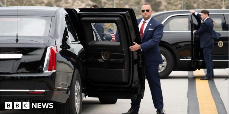 Two US Secret Service agents, both in suits, one in sunglasses, hold open the back passenger doors on two versions of the "Beast" armoured limousine.