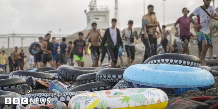 Rubber flotation rings sit on a beach in front of a line of arriving migrants in Ceuta.