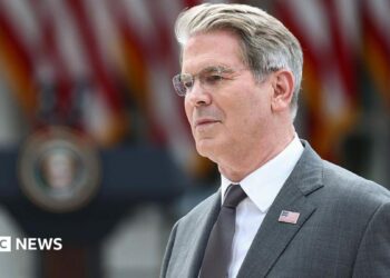 US Treasury Secretary Scott Bessent's head and shoulders in profile. He has grey hair and glasses. He is wearing a grey suit with a US flag lapel pin, a silver tie, and a white shirt. Behind him in soft focus are US flags and a podium.