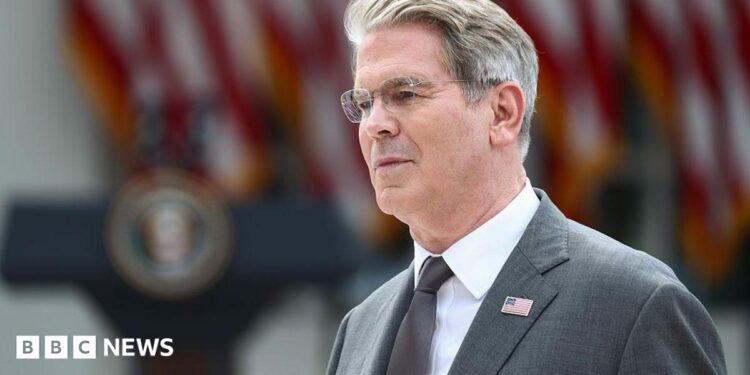 US Treasury Secretary Scott Bessent's head and shoulders in profile. He has grey hair and glasses. He is wearing a grey suit with a US flag lapel pin, a silver tie, and a white shirt. Behind him in soft focus are US flags and a podium.