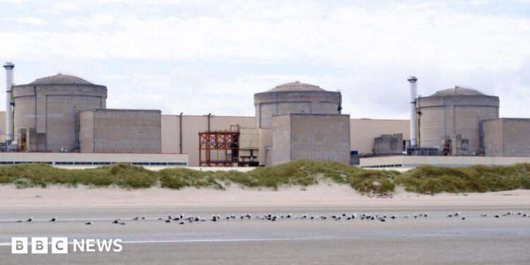A still image of the Gravelines nuclear power plant, with three stone-coloured units connected by a large white wall, set behind a stretch of empty coast with the tide out.